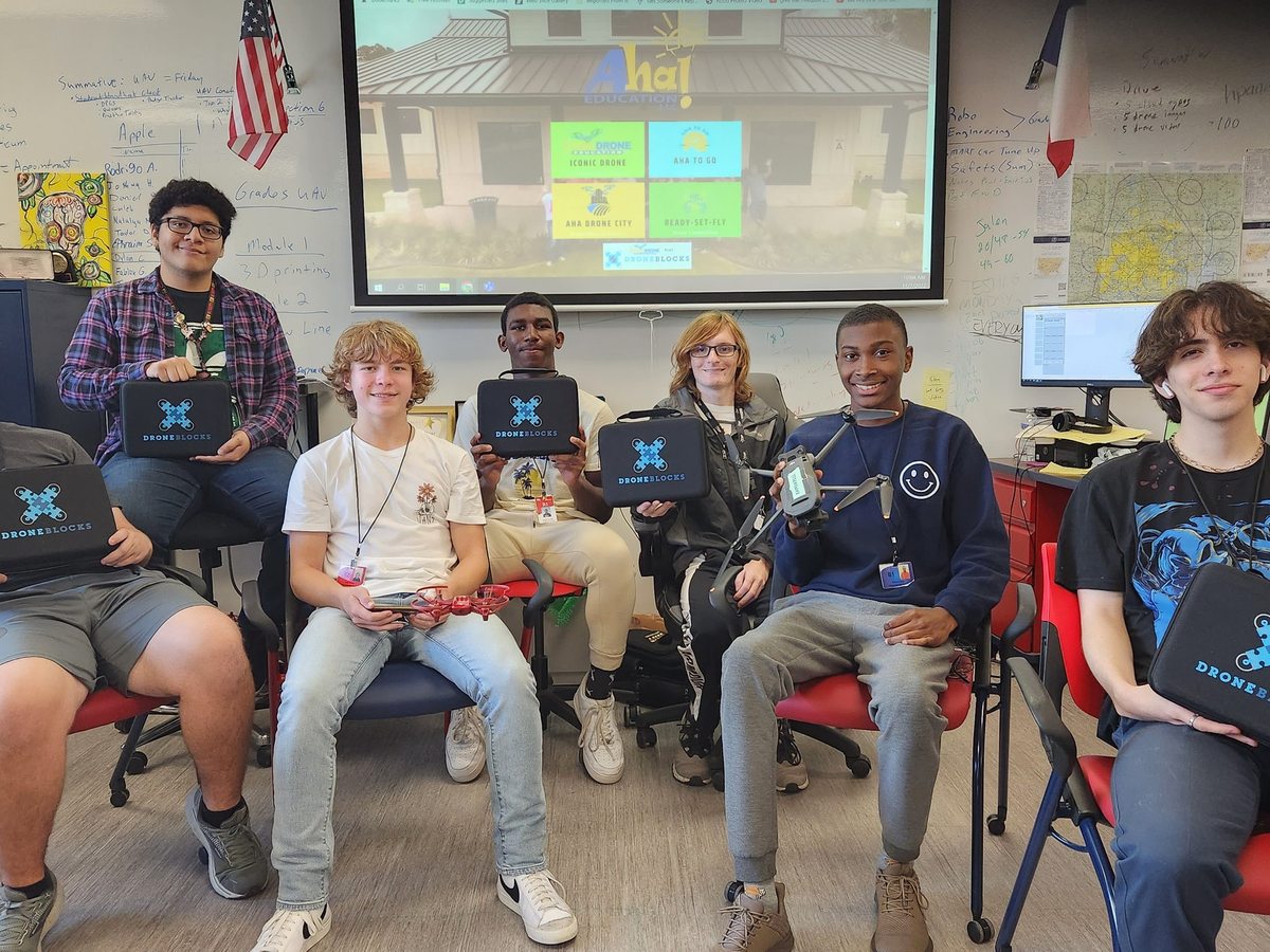 Students in a classroom holding laptop cases branded DroneBlocks, a lesson on the screen behind them.