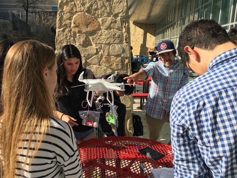 Students gathered outdoors around a drone, one holding it up as the others watch.
