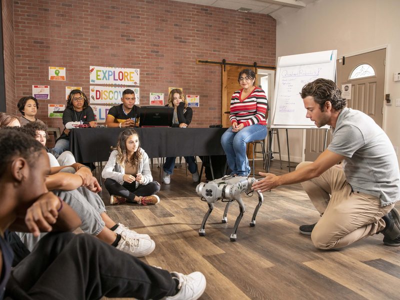A presenter kneeling with a quadruped robot in front of a seated audience.