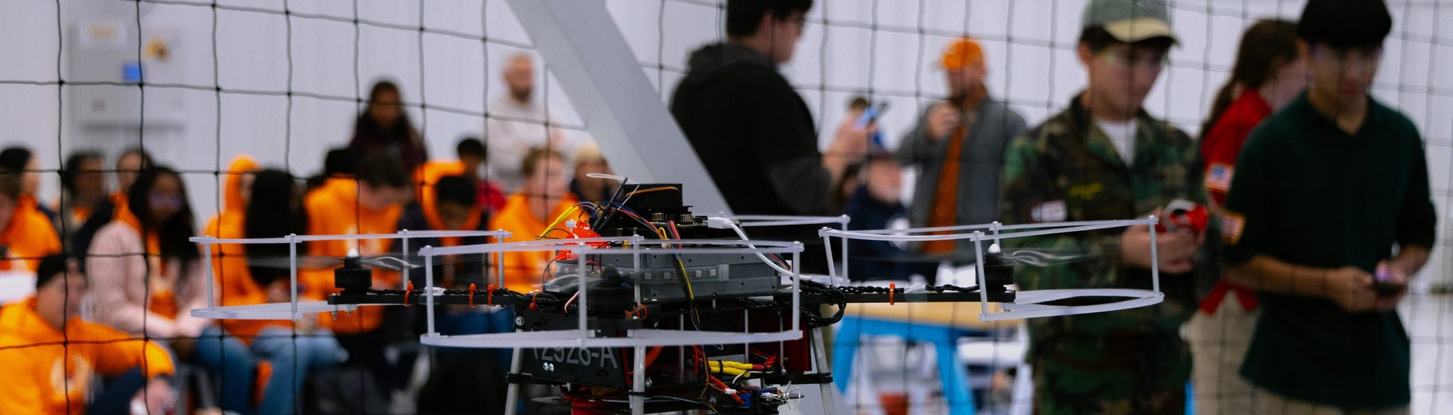 A competition drone airborne inside a netted arena, students and officials watching from the side.