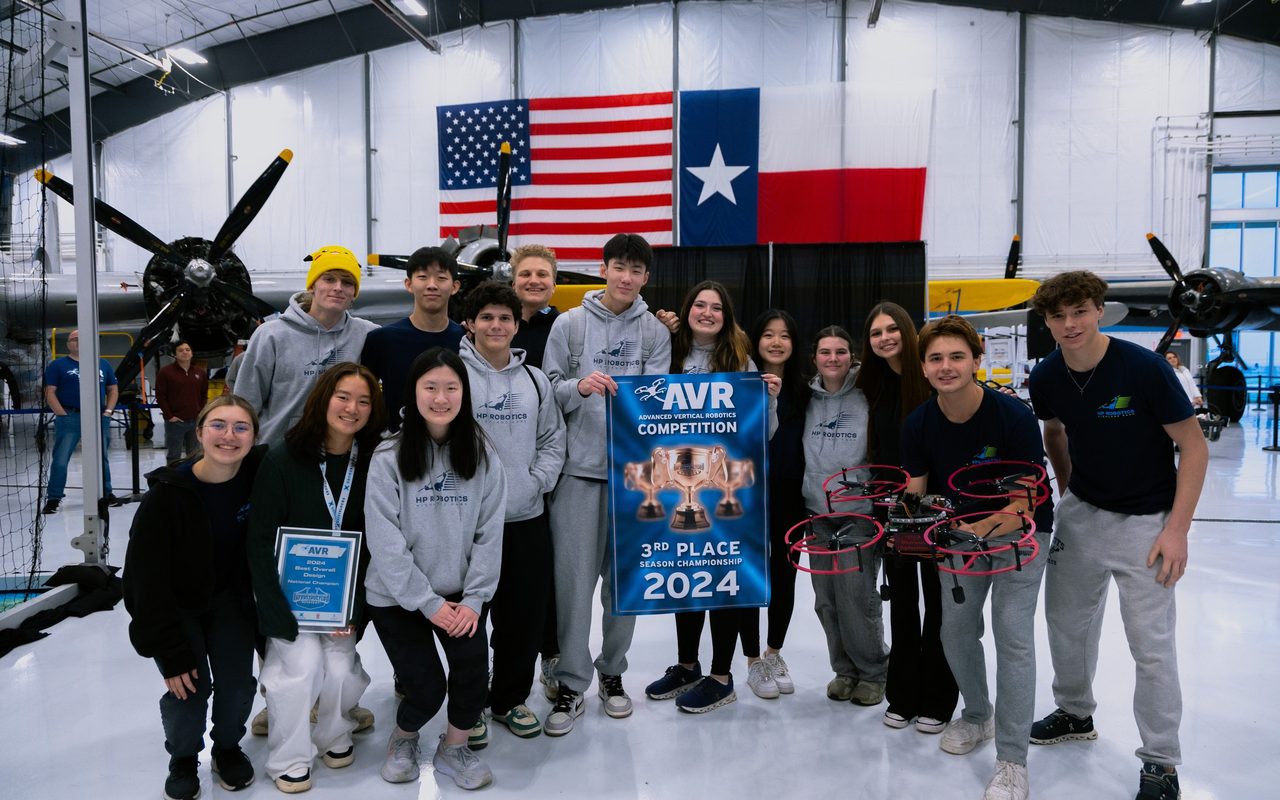 A team holding their third place banner and plaque beside their drones, under the flags in the hangar.