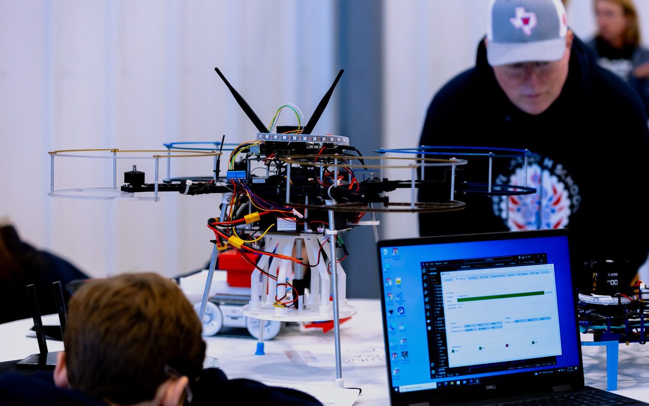 A large competition drone on a bench beside a laptop showing telemetry, students and a mentor working behind it.
