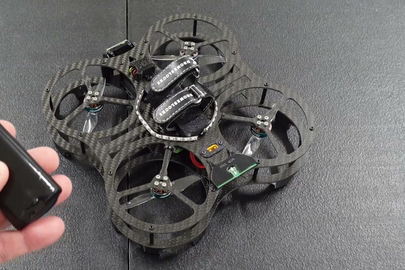 Close view of a DEXI-3 on the bench before a flight: carbon-fibre ducted frame, four motors, LED ring and flight controller visible, with a hand holding the battery about to go in.