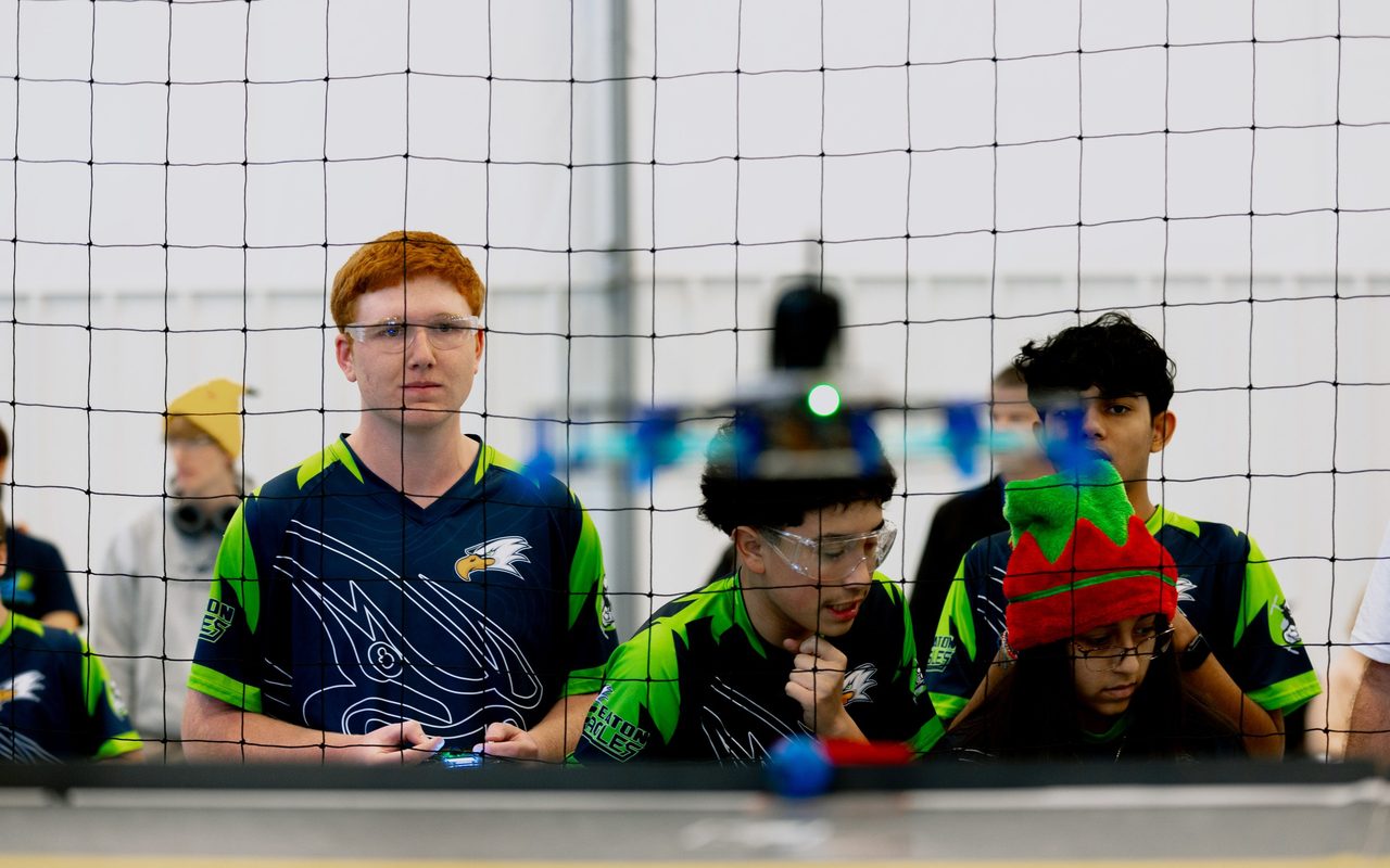 Four students in team jerseys watching from behind the safety netting, a drone in the air beyond.