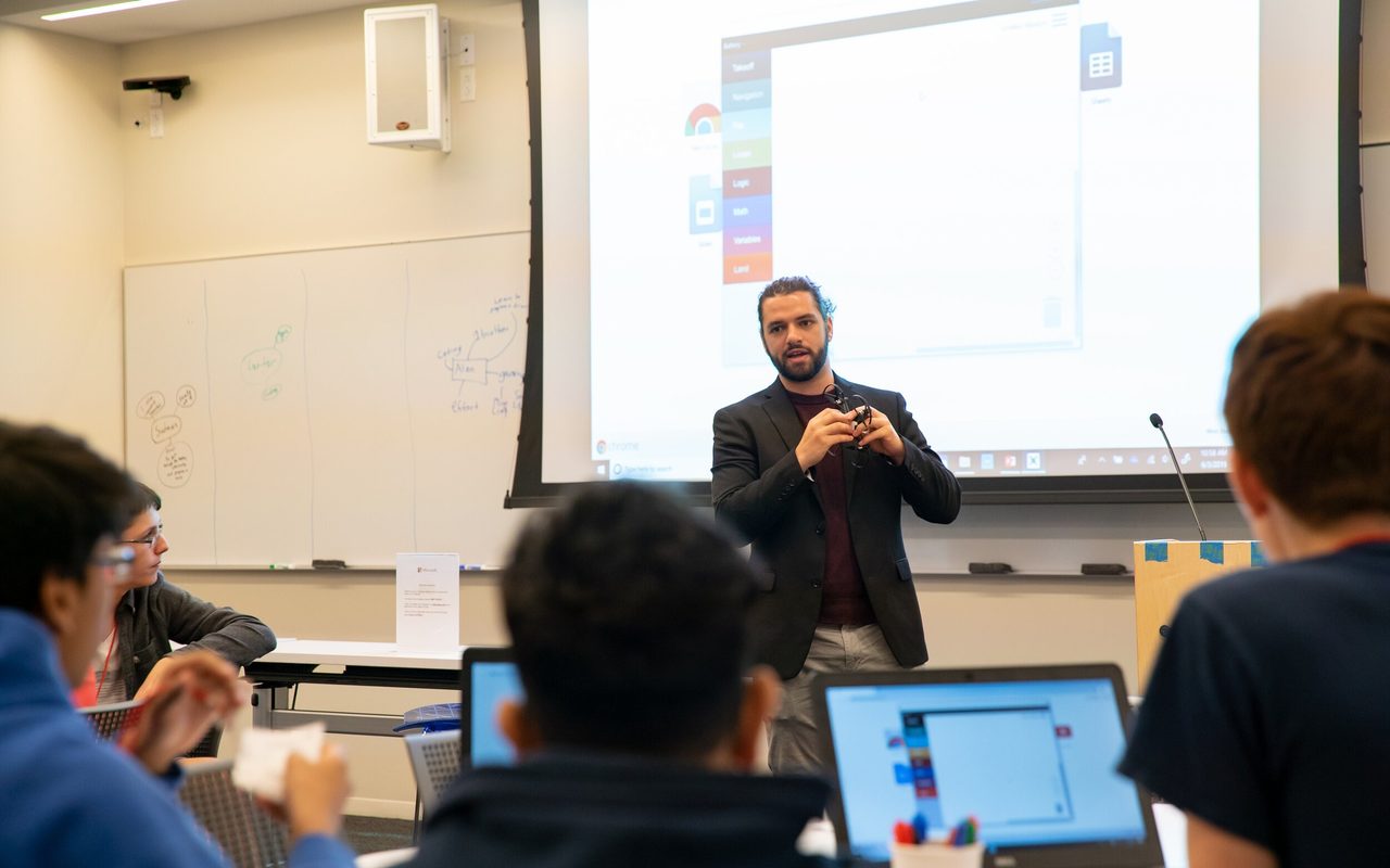 An instructor presenting to a seated class with laptops and tablets open.