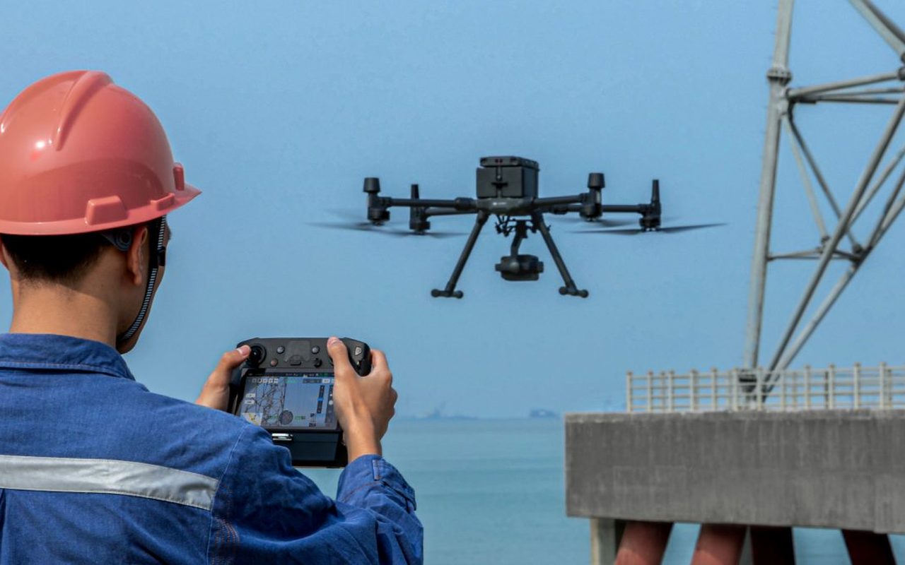 A worker in a hard hat flying an inspection drone beside industrial structures.
