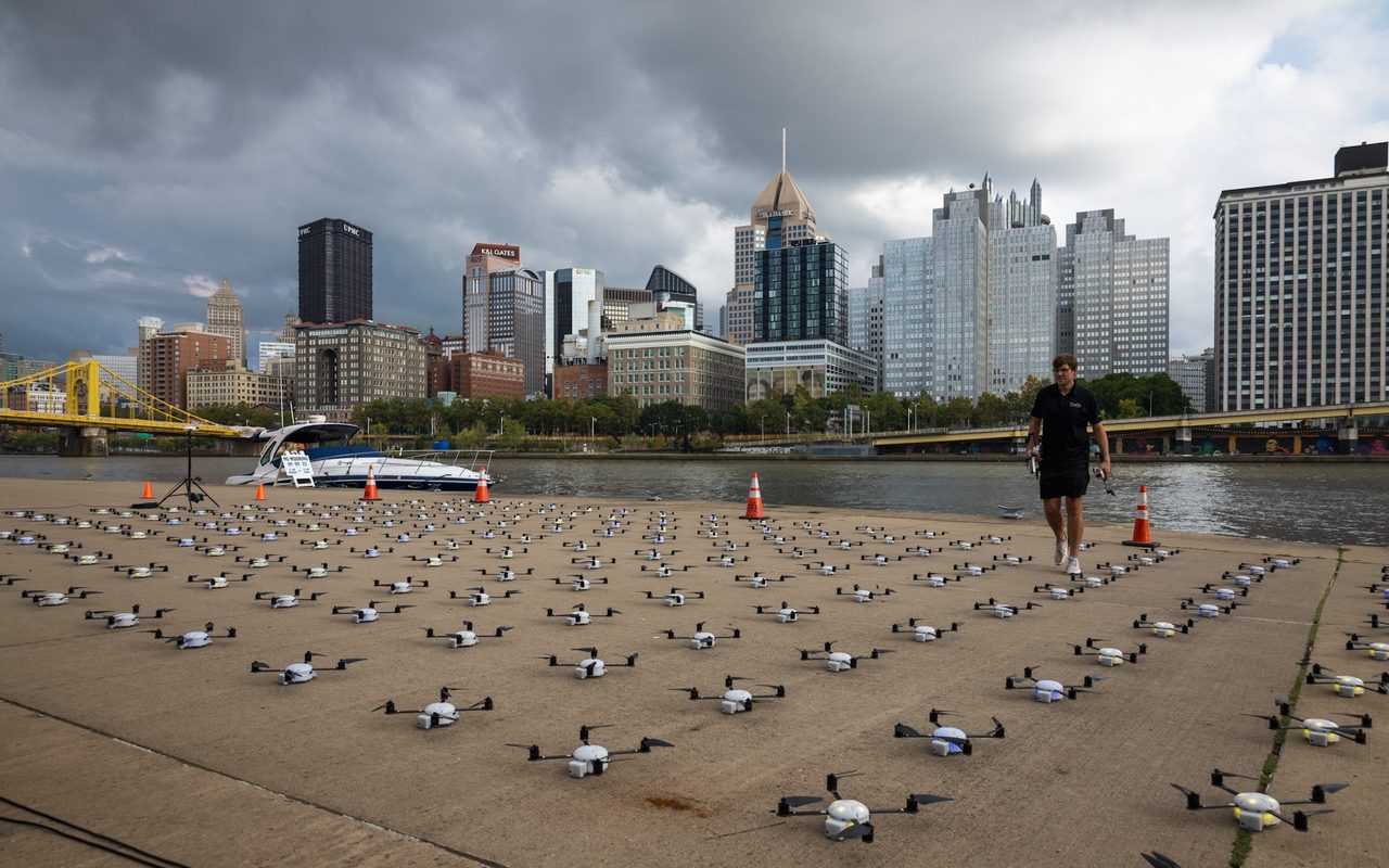 A fleet of drones laid out in a grid on a riverside plaza before a light show, city skyline behind.